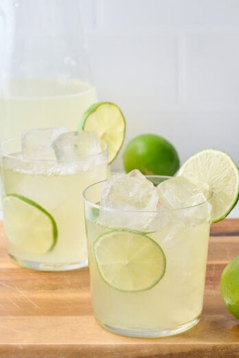 Two glasses of homemade limeade with lime slices tucked into glass and on rim with extra limes around and pitcher nearby.