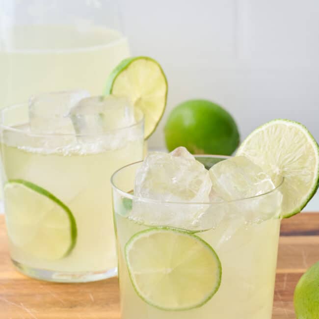 Two glasses of homemade limeade with lime slices tucked into glass and on rim with extra limes around and pitcher nearby.