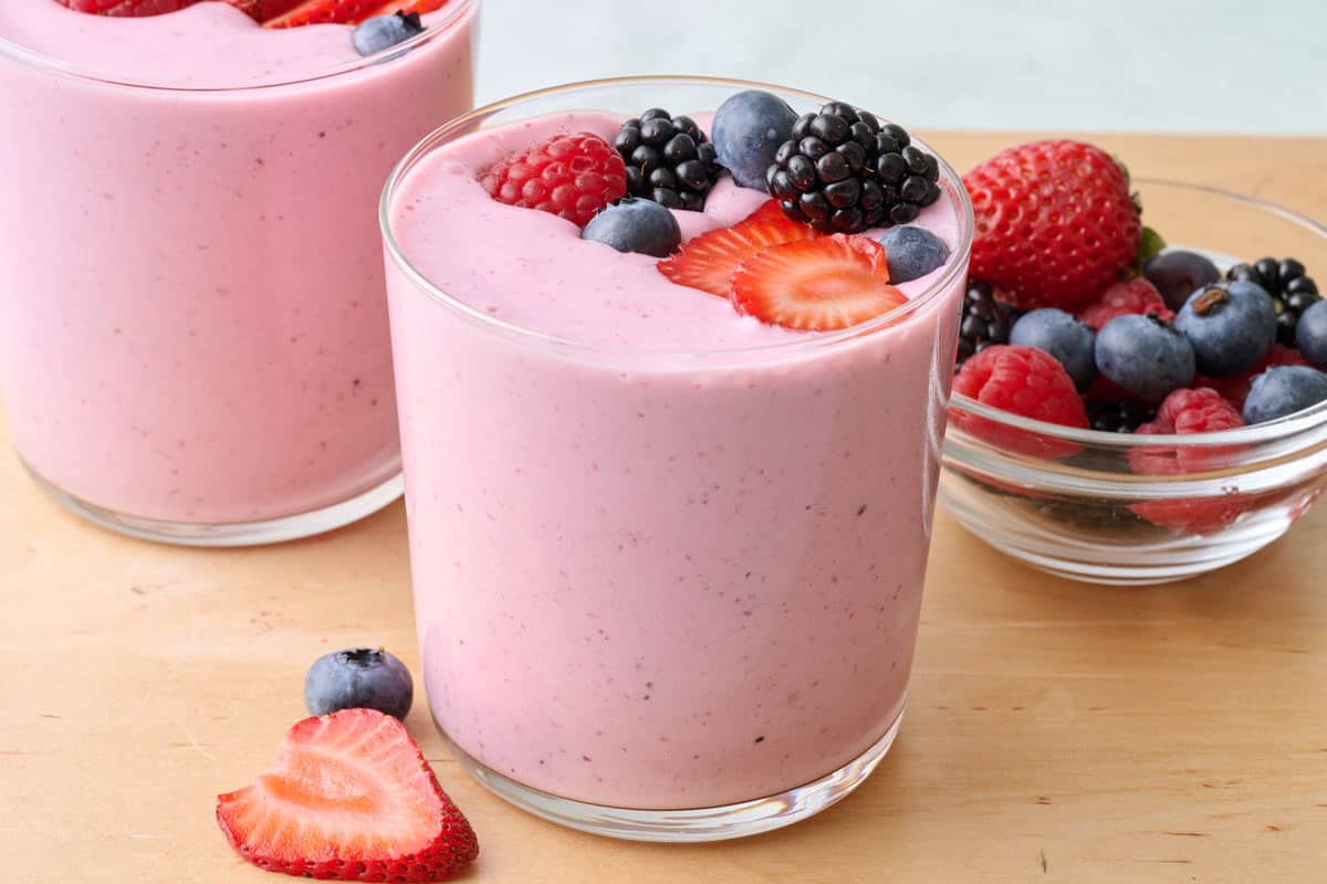 2 glasses of mixed berry smoothie topped with extra mixed berries and more berries either in a dish nearby.
