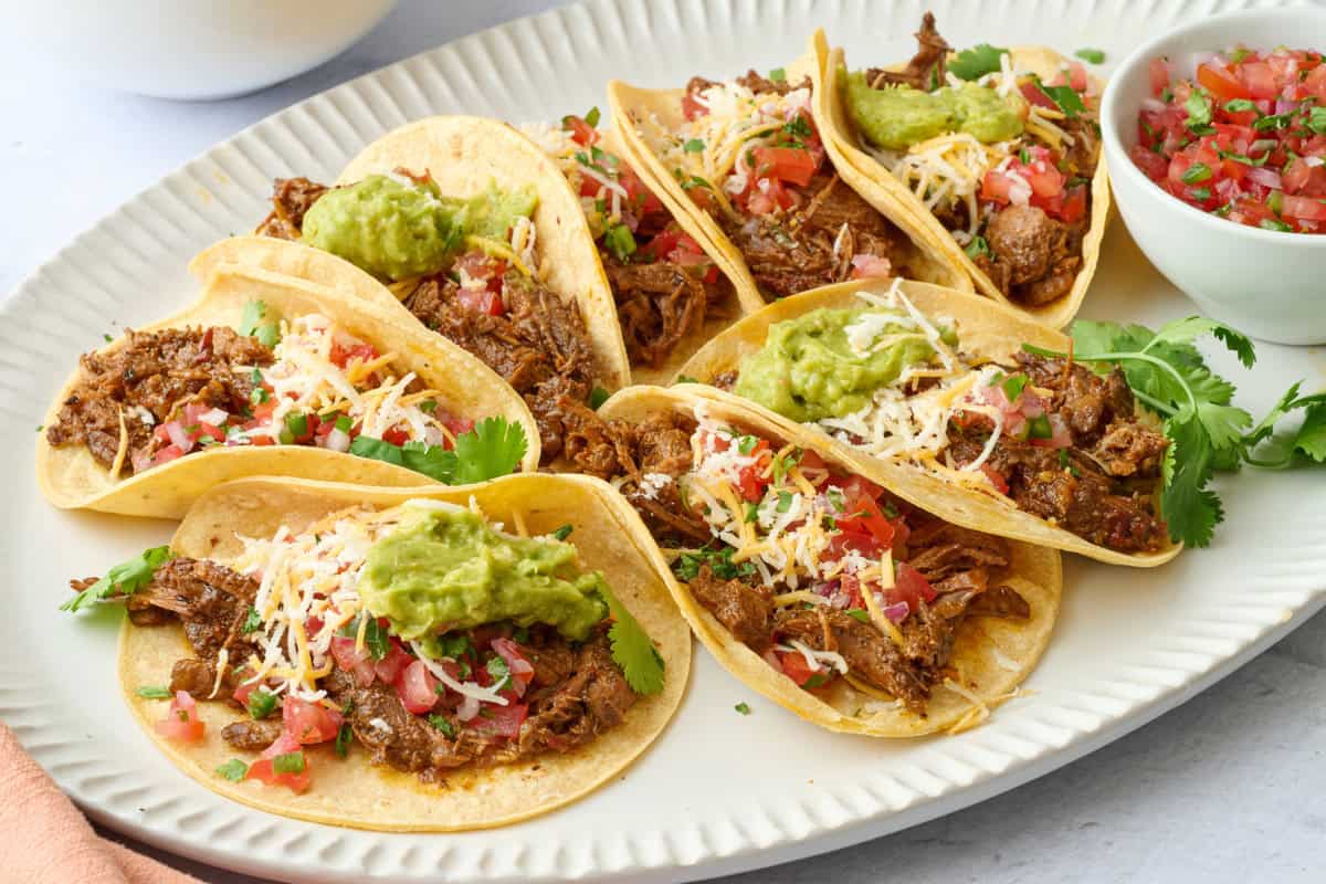 Shredded beef tacos on a platter topped with fresh pico de gallo, guacamole, shredded cheese.