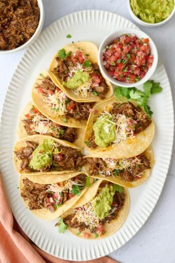 Shredded beef tacos on a platter topped with fresh pico de gallo, guacamole, shredded cheese.