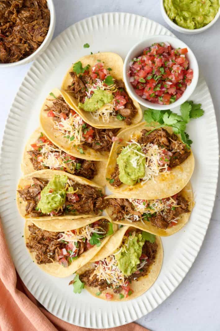 Shredded beef tacos on a platter topped with fresh pico de gallo, guacamole, shredded cheese.