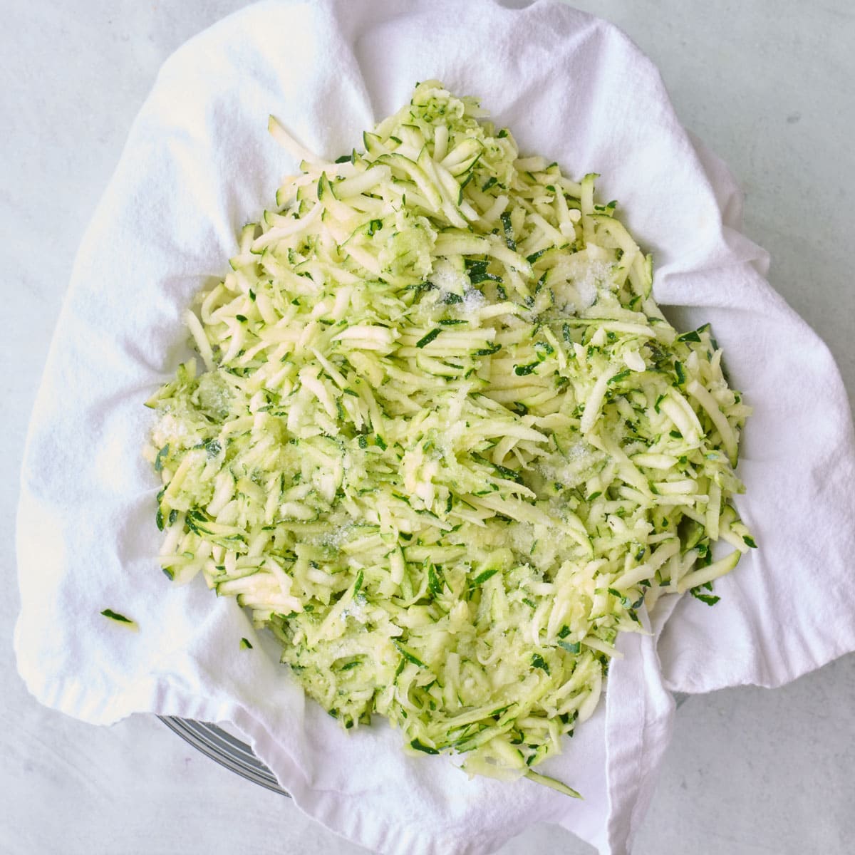 Grated zucchini in a kitchen towel with salt sprinkled on top in a fine mesh sieve.
