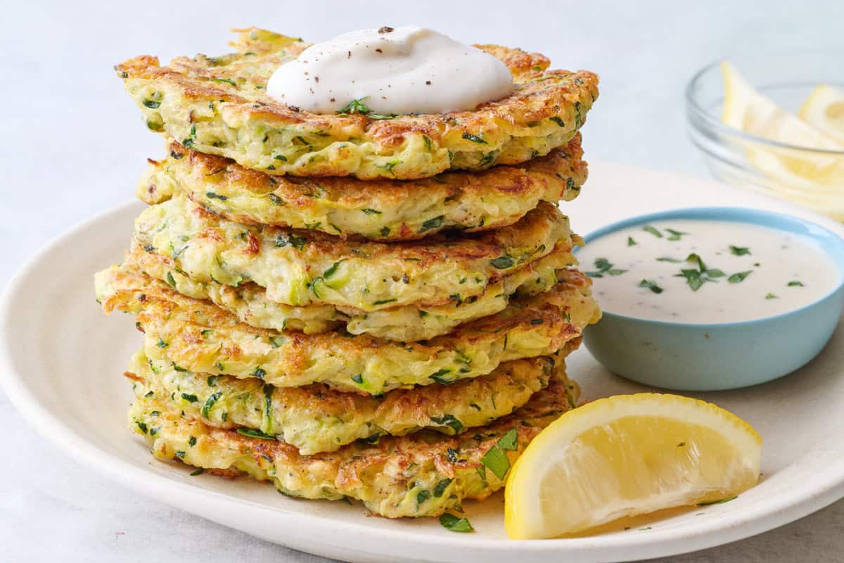 Easy zucchini fritters recipe stacked up on a plate with a dollop of ranch on top and a small dish of more nearby.