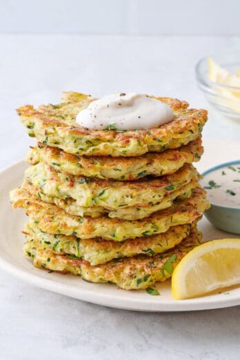 Easy zucchini fritters recipe stacked up on a plate with a dollop of ranch on top and a small dish of more nearby.