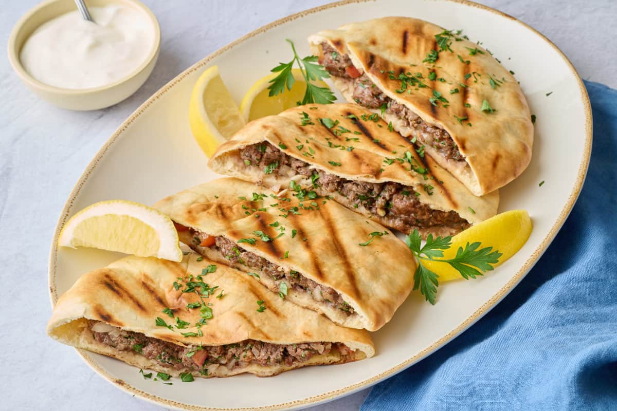 Pan grilled pitas cut into halves, layered on a narrow platter with a side of yogurt with spoon dipped inside, lemon wedges and parsley.