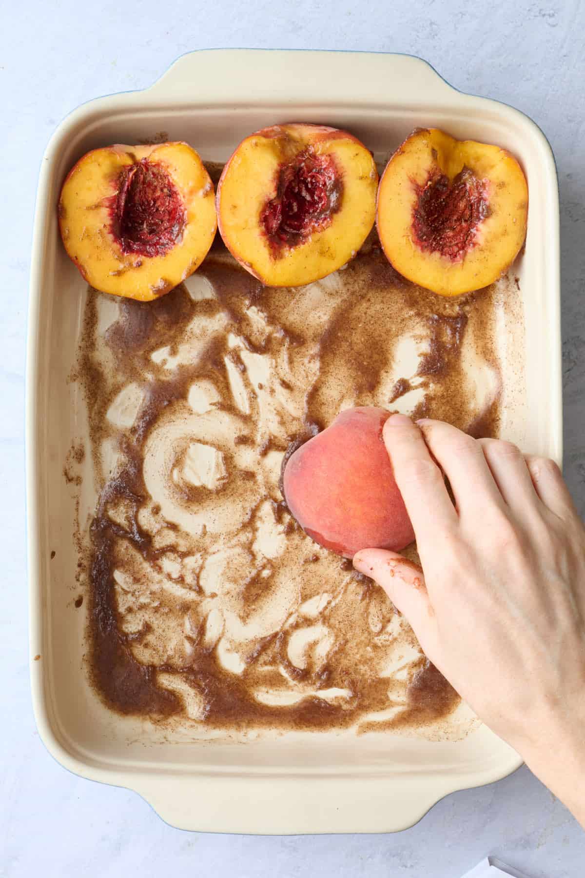 A few peaches in a baking dish with one cut side down and a hand rubbing the spiced butter on to it.