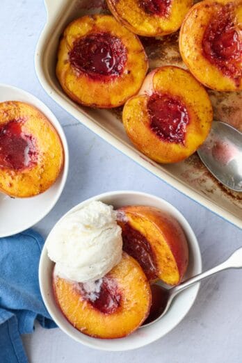 Small bowl with two baked peach halves topped with ice cream, a baking dish of more roasted peaches nearby.