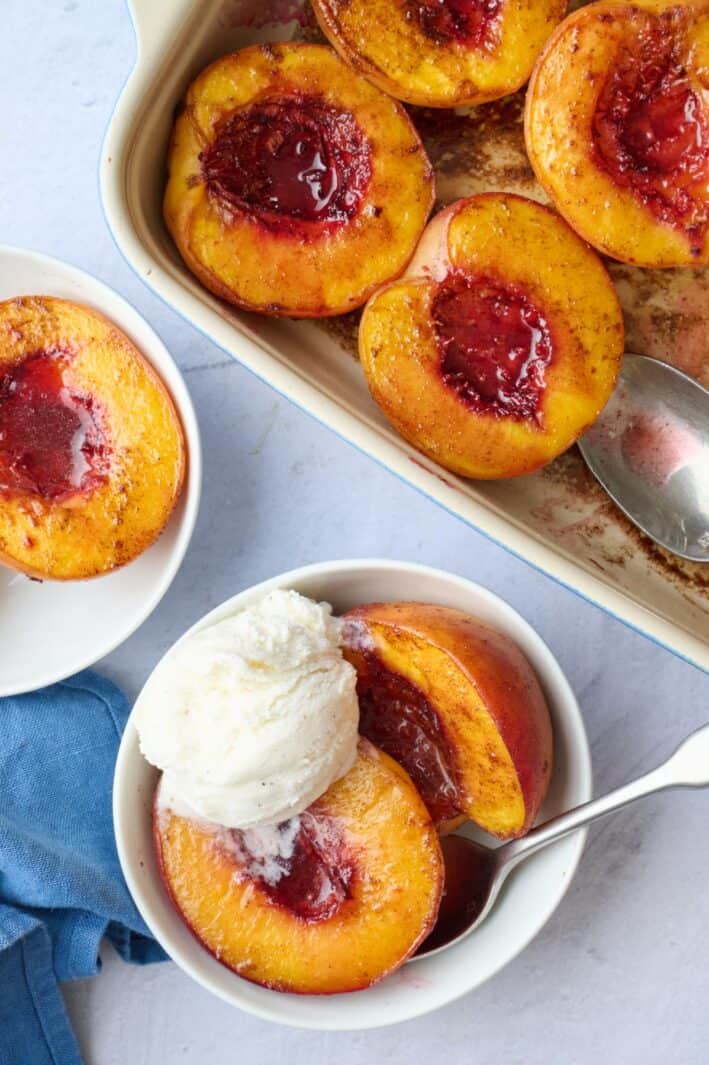 Small bowl with two baked peach halves topped with ice cream, a baking dish of more roasted peaches nearby.