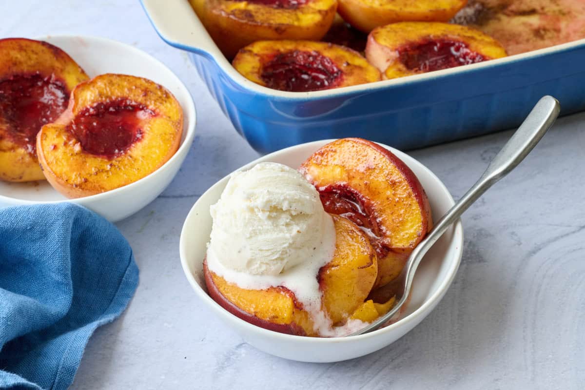 Two servings of homemade baked peaches in bowls, one topped with ice cream and baking dish of more nearby.