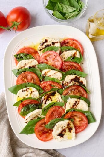 Assembled caprese salad on an oval platter, seasoned with flaky salt and pepper and a drizzle of oil and balsamic glaze on top.