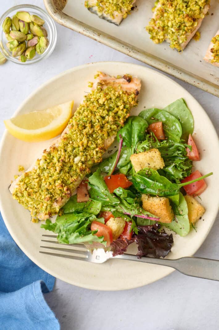 Pistachio crusted salmon fillet on a plate with a light spring salad, sheet pan nearby.