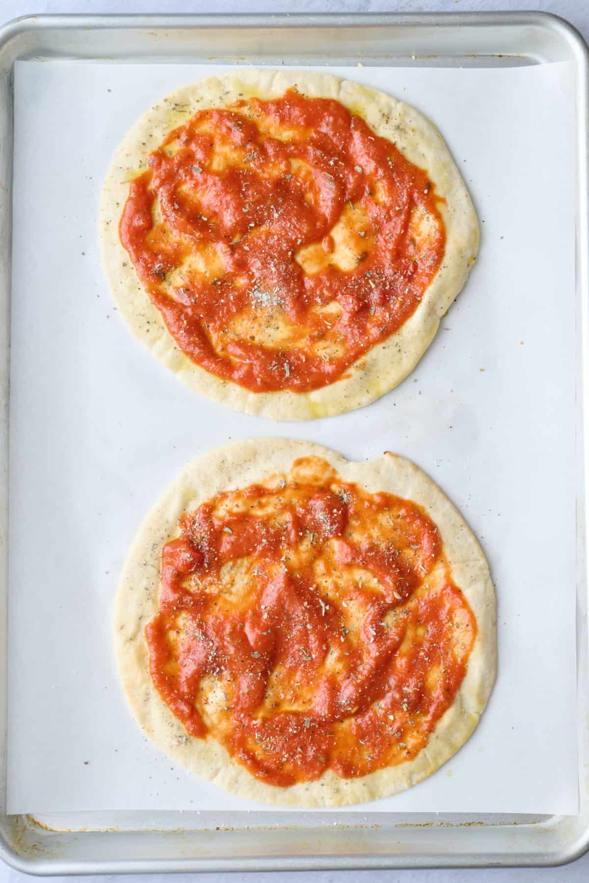 Two pitas on a sheet pan with marinara sauce and seasonings sprinkled on top.