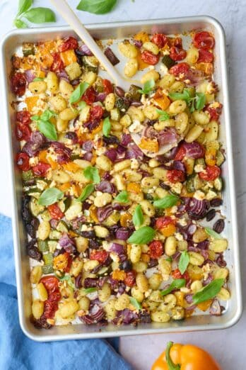 Sheet pan gnocchi in pan with a spatula under some, garnished with fresh basil, extra fresh ingredients around.