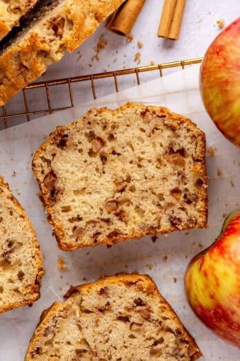 Slice of homemade apple bread on parchment paper with more slices around and a few whole honey crisp apples and cinnamon sticks nearby.