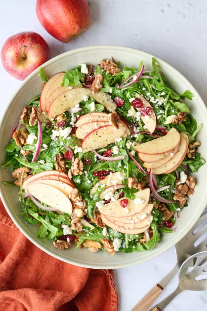 Apple walnut salad in a serving bowl topped with a few bunches of fanned out apples slices, and extra toppings, salad serving set resting nearby and a fresh apple peaking from corner.