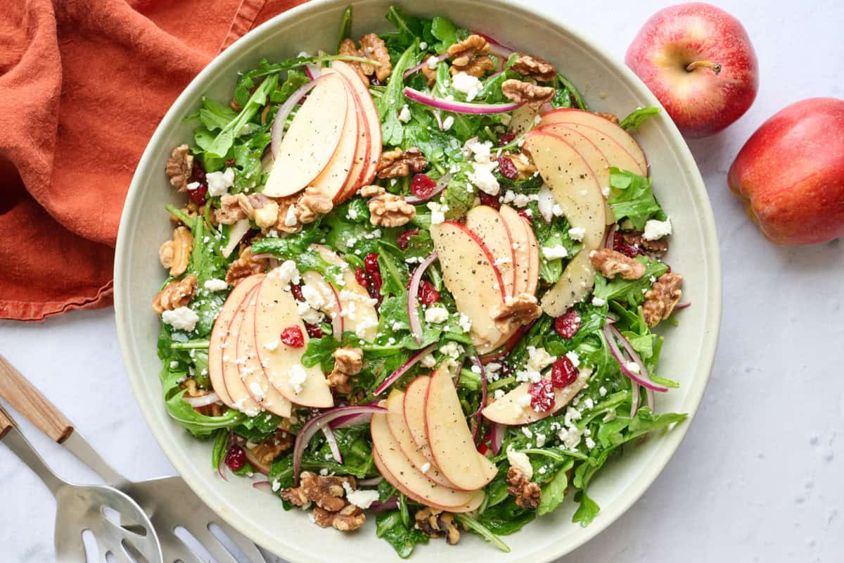 Apple walnut salad in a serving bowl topped with a few bunches of fanned out apples slices, and extra toppings, salad serving set resting nearby and a fresh apple peaking from corner.