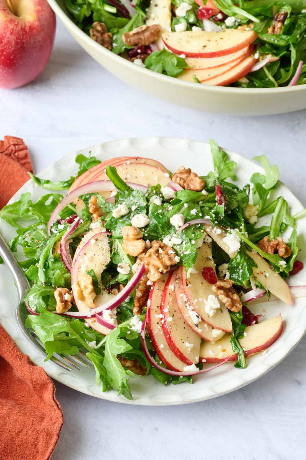 Serving of apple walnut salad on a small plate with fanned out apples, fork resting on plate, and salad bowl nearby.