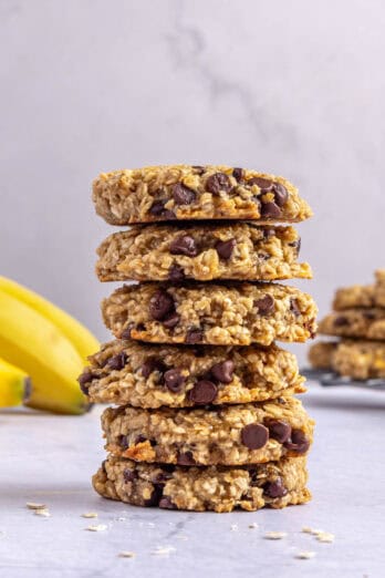 Banana oatmeal Cookies stacked up with more around, wire rack and a few whole bananas in the background.