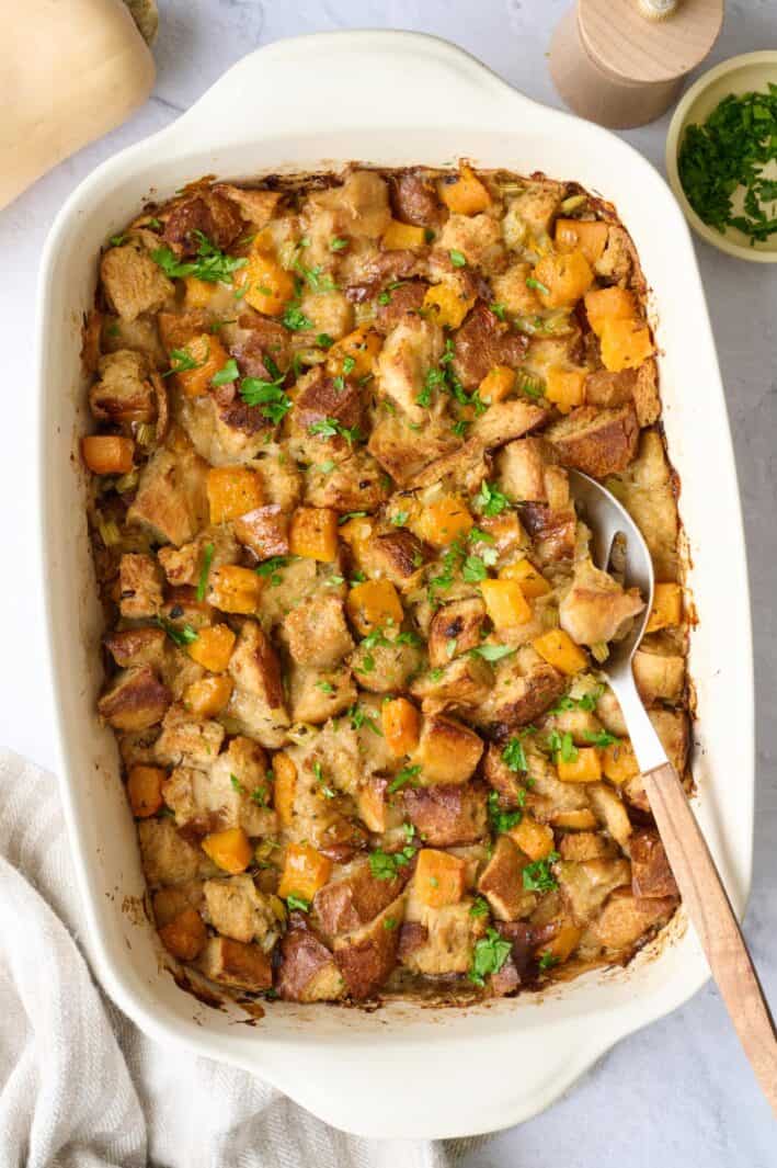 Butternut squash stuffing in baking dish, garnished with fresh parsley, serving spoon dipped inside.
