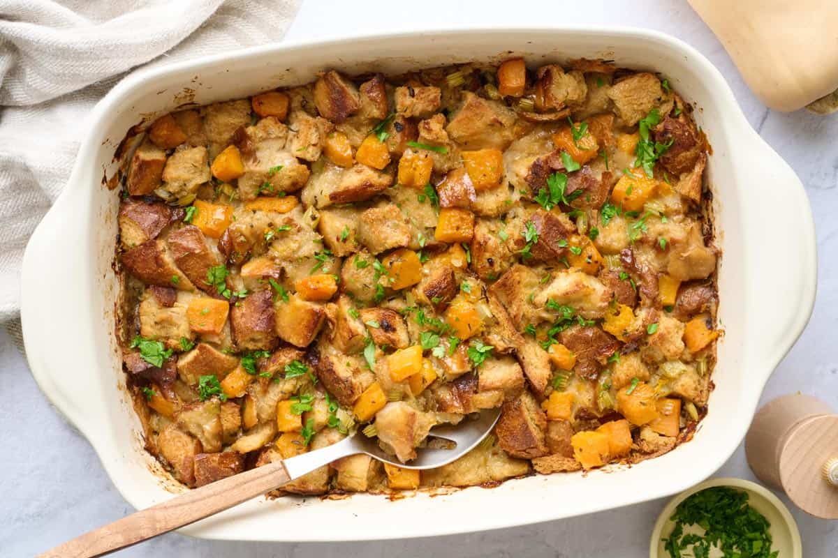 Butternut squash stuffing in baking dish, garnished with fresh parsley, serving spoon dipped inside.