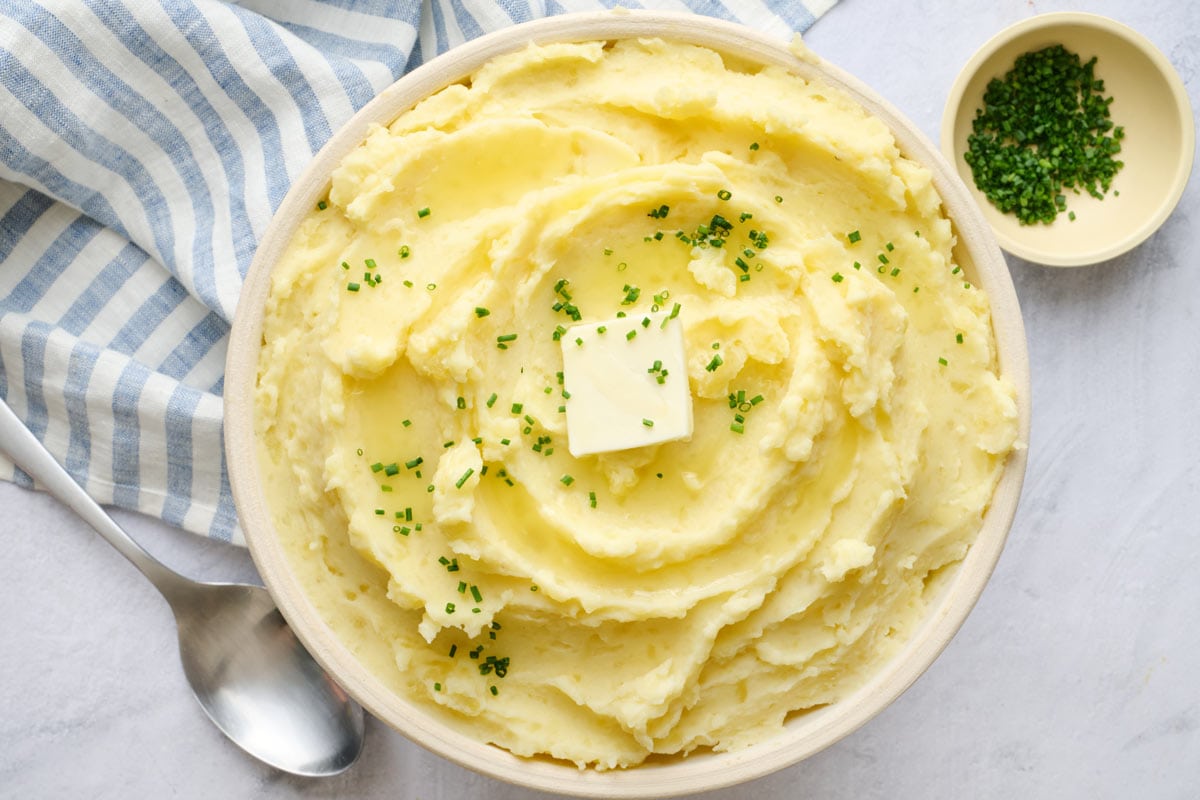 Creamy mashed potatoes in a serving bowl with a pat a mostly melted butter on top, garnished with chopped chives, serving spoon nearby.