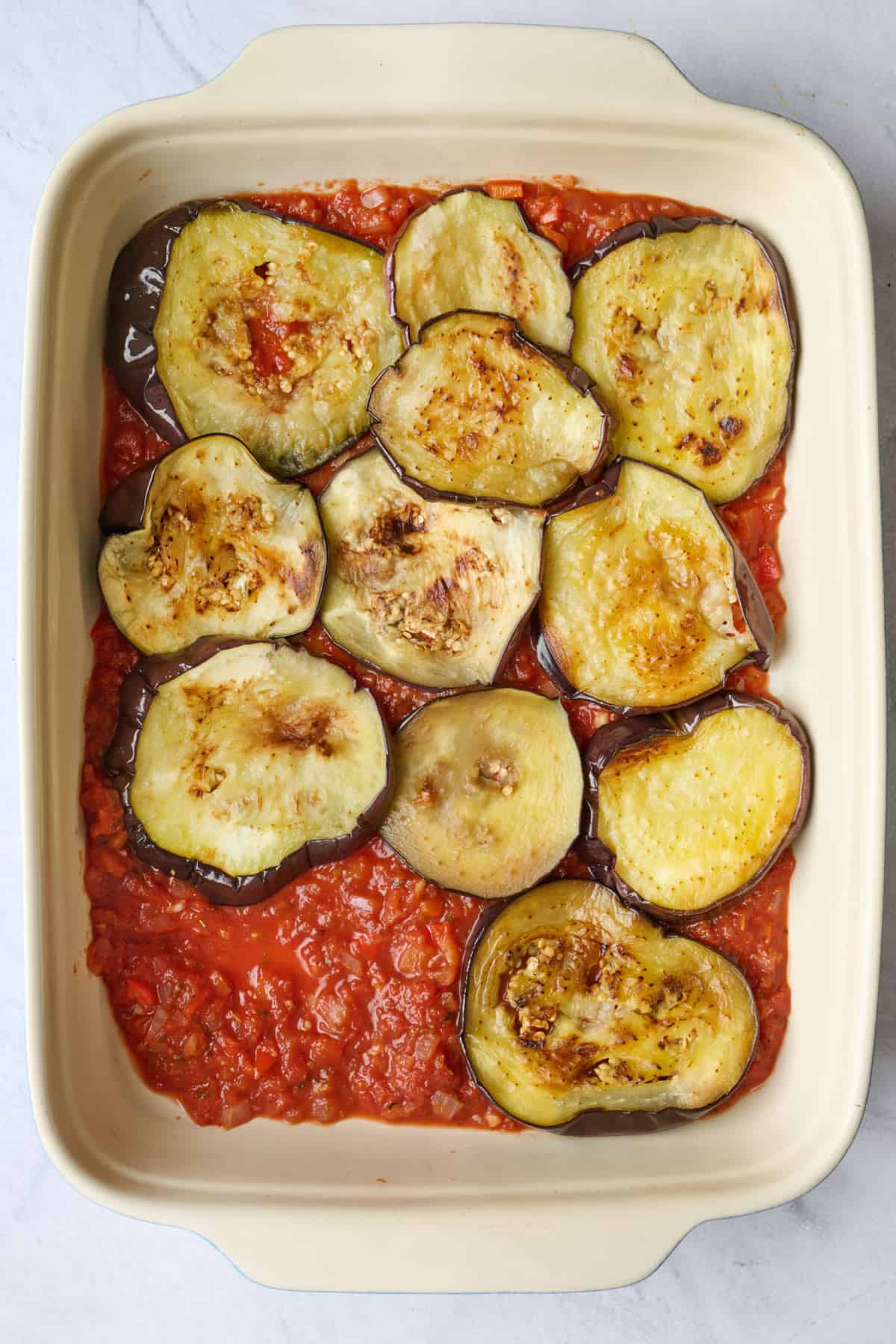 Tomato sauce spread in a thin layer of baking dish with half of the eggplant slices layered into dish.