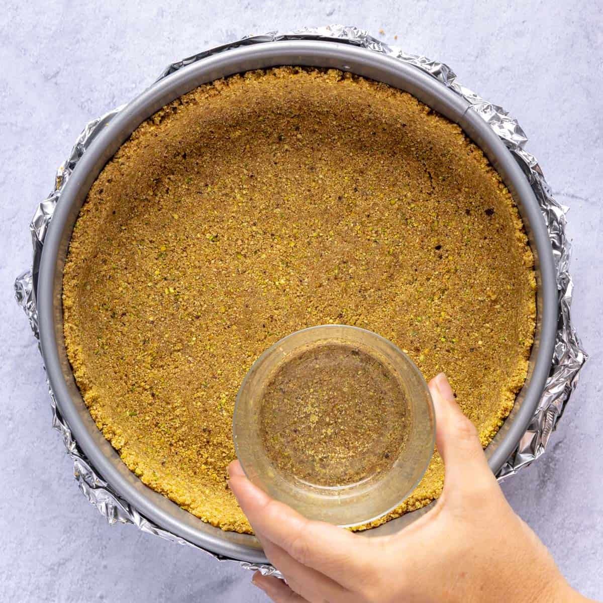 Hand using a glass to press the graham cracker crumbs into springform pan.