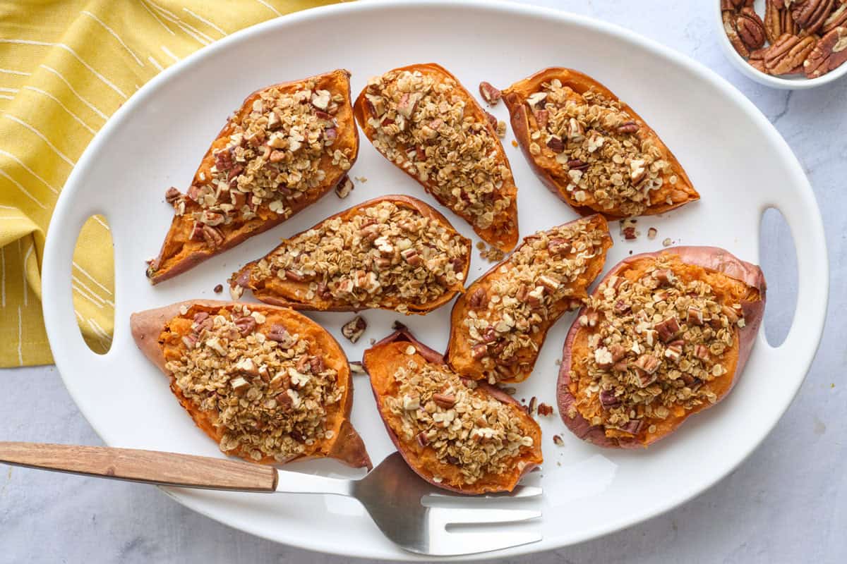 Twice baked sweet potatoes halves on a narrow platter with serving fork and small dish of chopped pecans.