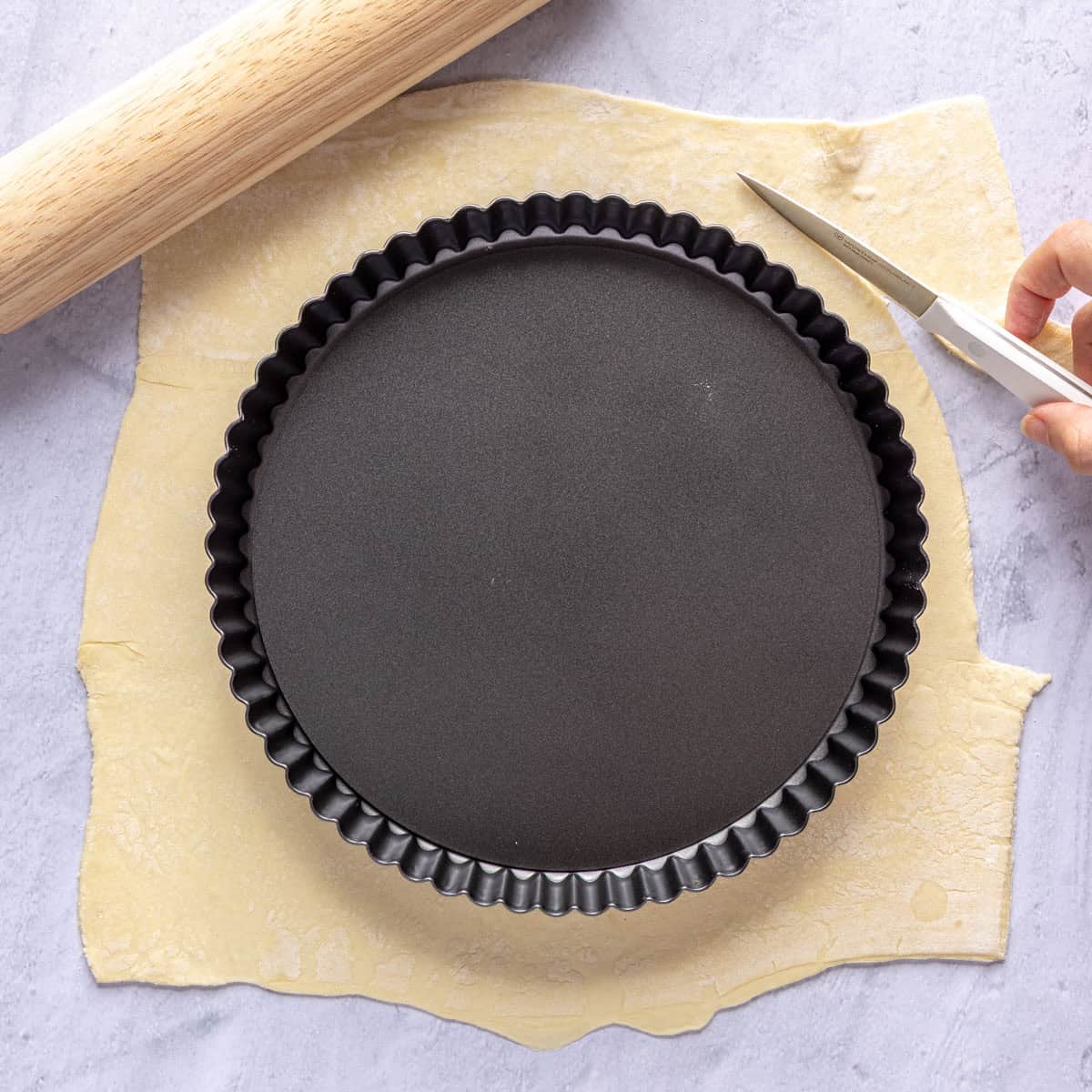 Puff pastry rolled out with a tart pan on top and a knife cutting a circle with enough pastry to go up sides of pan.