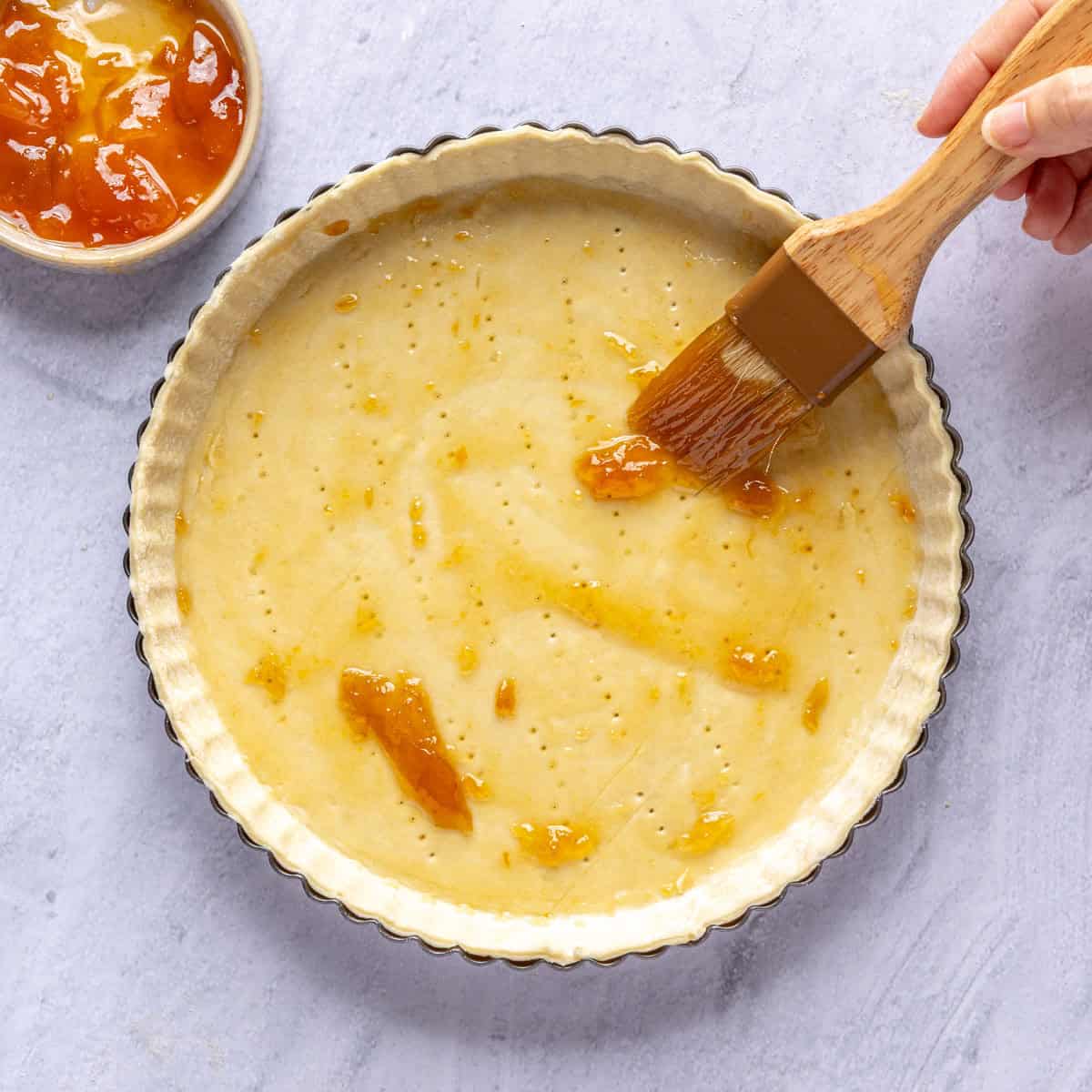 Puff pastry pressed into a tart pan with a pastry brush adding apricot jam to the bottom.