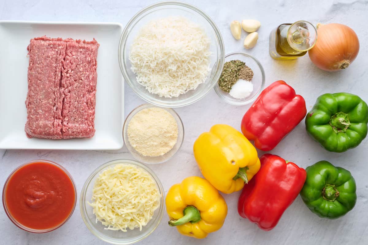 Ingredients for recipe before prepping: lean ground beef, bell peppers, tomato sauce, shredded mozzarella cheese, grated parmesan cheese, cooked white rice, seasonings, garlic cloves, onion, and olive oil.