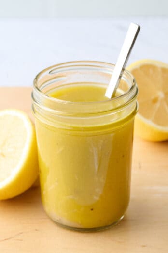 Easy lemon vinaigrette dressing in a small mason jar with a spoon dipped inside and a few fresh lemon halves nearby.