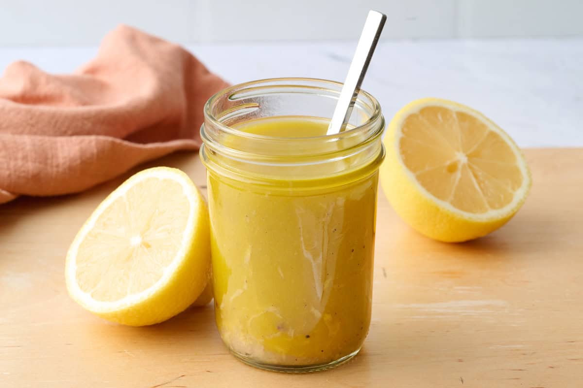 Quick homemade lemon vinaigrette in a small jar with a spoon dipped in with fresh lemon nearby.