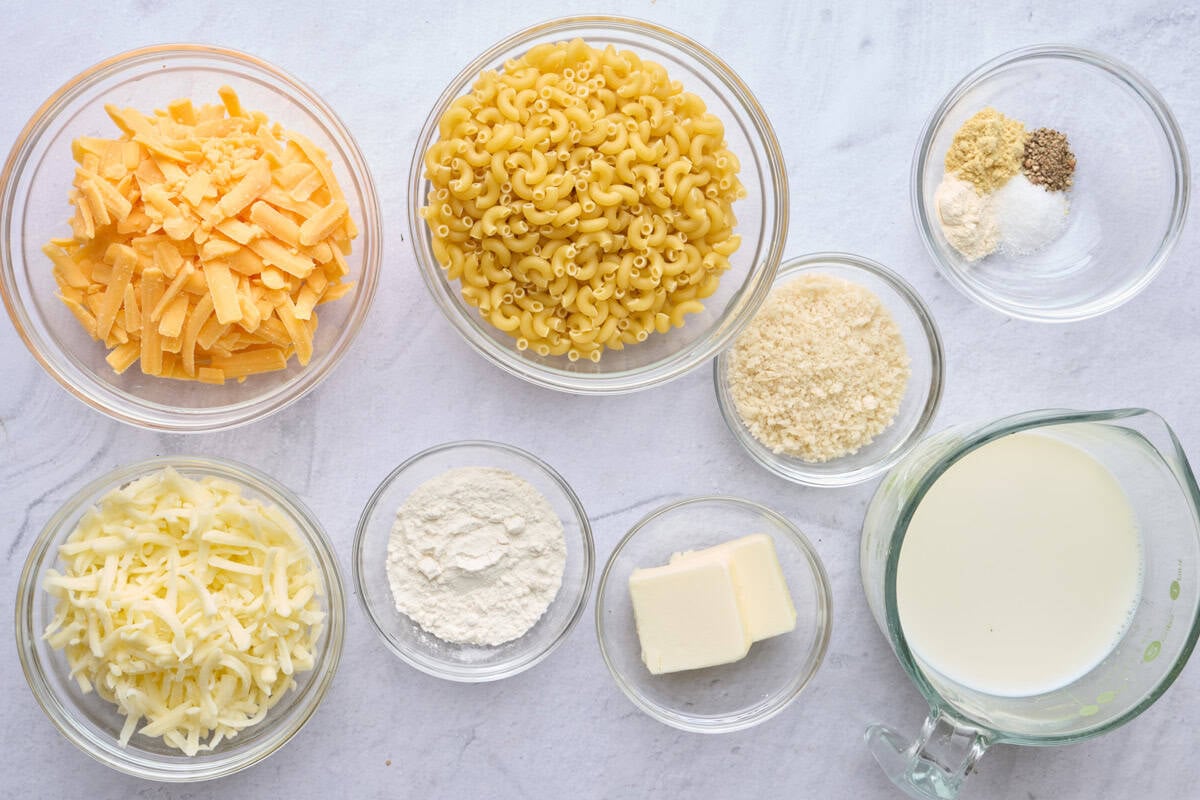 Ingredients for mini macaroni and cheese recipe in individual dishes: shredded cheddar cheese, shredded monterey jack cheese, elbow pasta, flour, breadcrumbs, butter, seasonings, and milk.