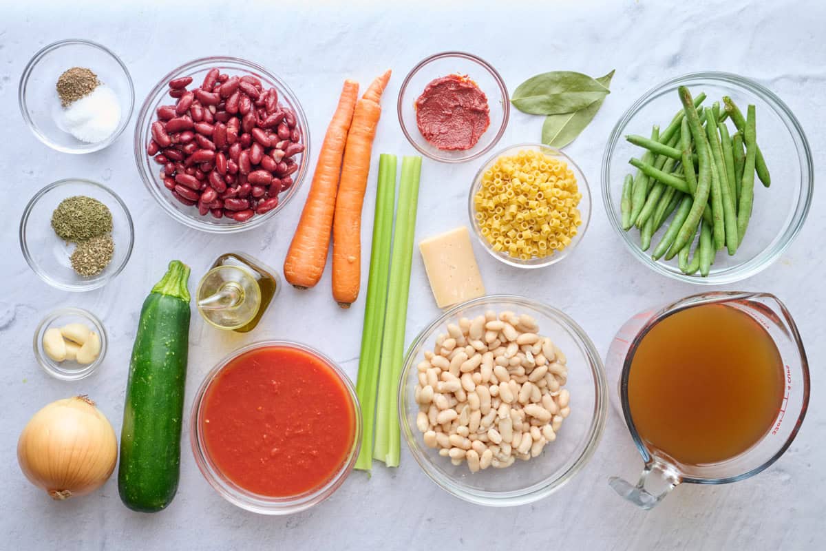 Ingredients for soup recipe: salt and pepper, dried herbs, garlic cloves, onion, zucchini, kidney beans, oil, tomato sauce, carrots, celery, tomato paste, pasta, parmesan rind, white beans, green beans, and broth.