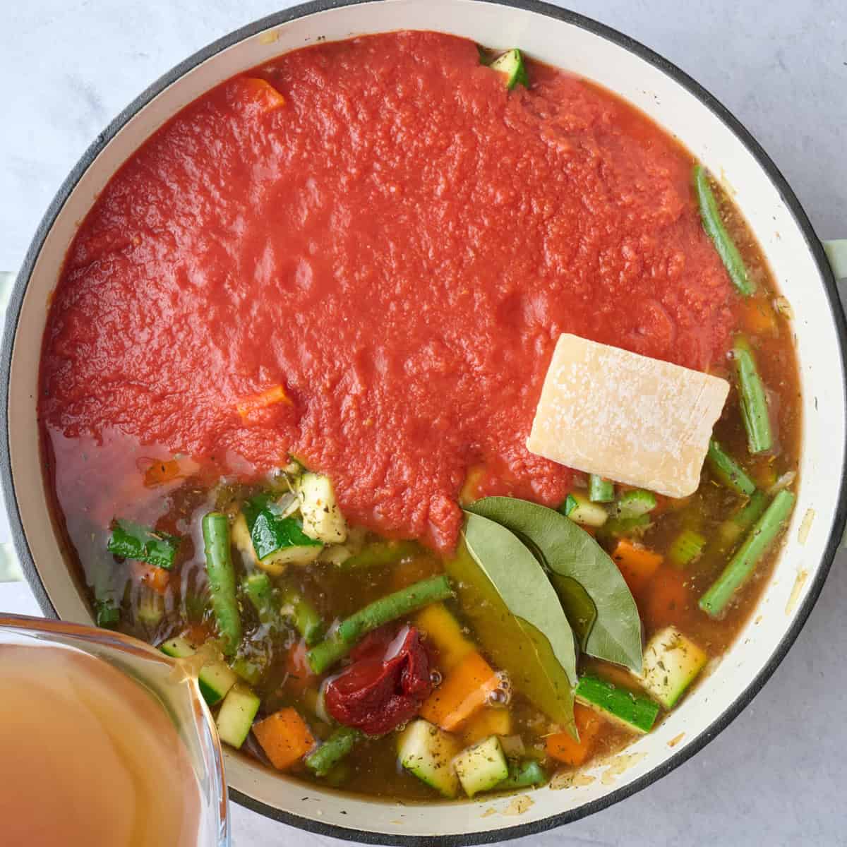 Broth being poured into the pot with tomato paste, crushed tomatoes, bay leaves, and parmesan rind before mixing.