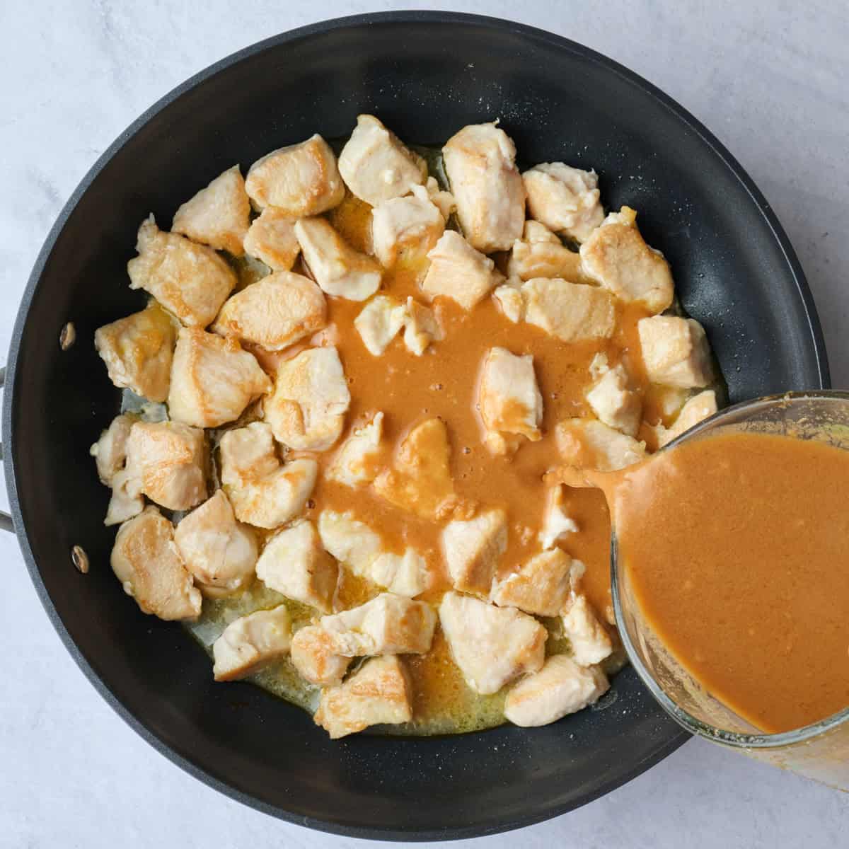 Savory peanut butter sauce being poured into skillet with cooked chicken pieces.