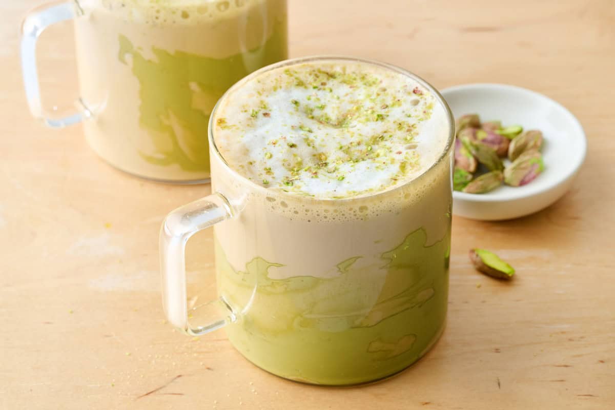Two mugs of hot pistachio latte using pistachio cream, garnished with frothed milk and crushed pistachios.
