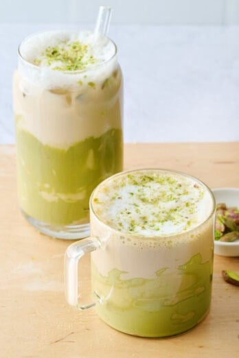 Hot pistachio latte in a glass mug with a tall glass of iced pistachio latte nearby.