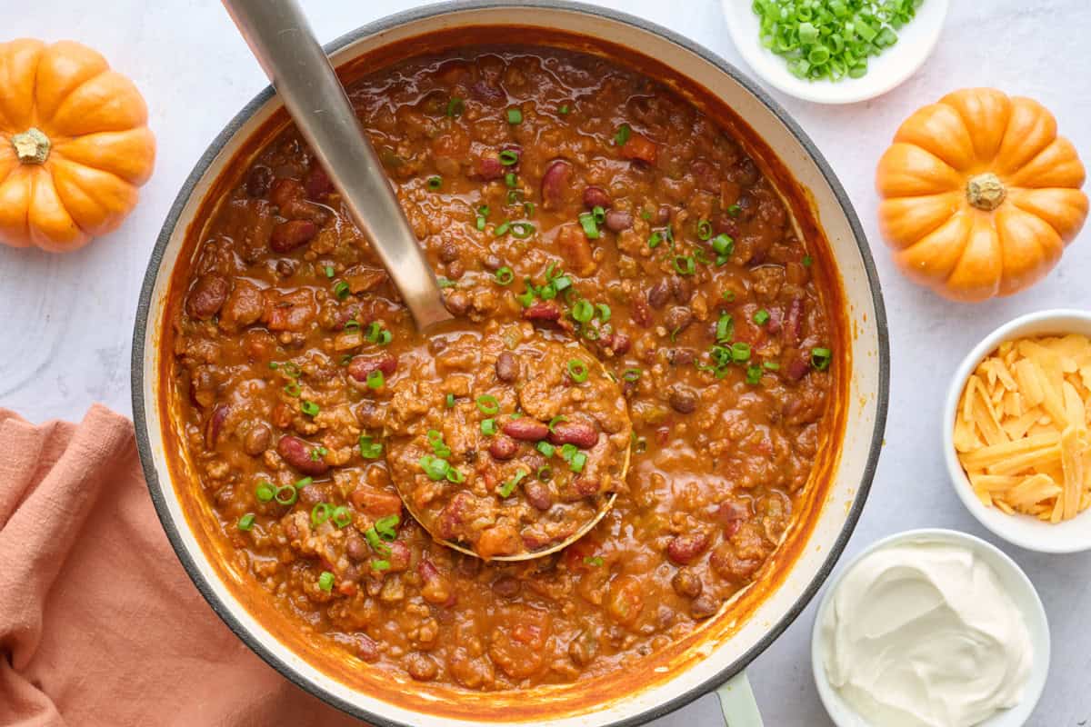 Pot of pumpkin black bean chili with shredded cheese, sour cream and chopped green onions on the side for toppings.