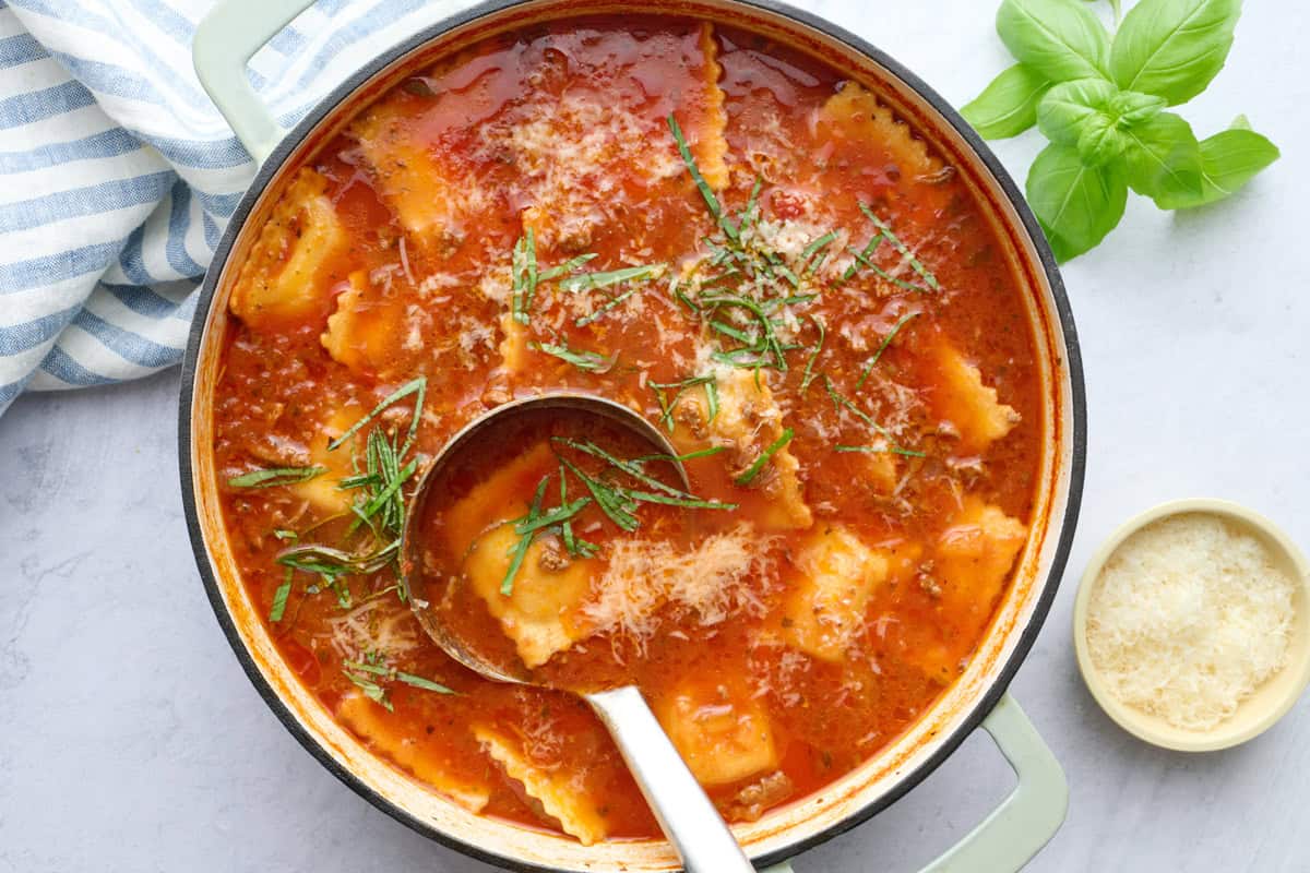 Ravioli soup recipe in a pot with a ladle dipped inside, garnished with parmesan cheese and sliced basil.