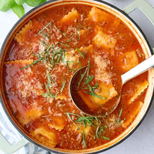 Ravioli soup recipe in a pot with a ladle dipped inside, garnished with parmesan cheese and sliced basil.