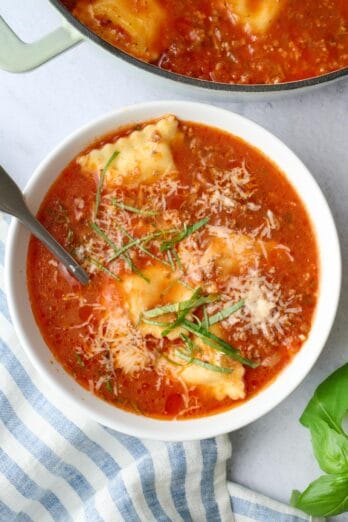 A bowl of beef ravioli soup garnished with grated parmesan cheese and fresh basil.