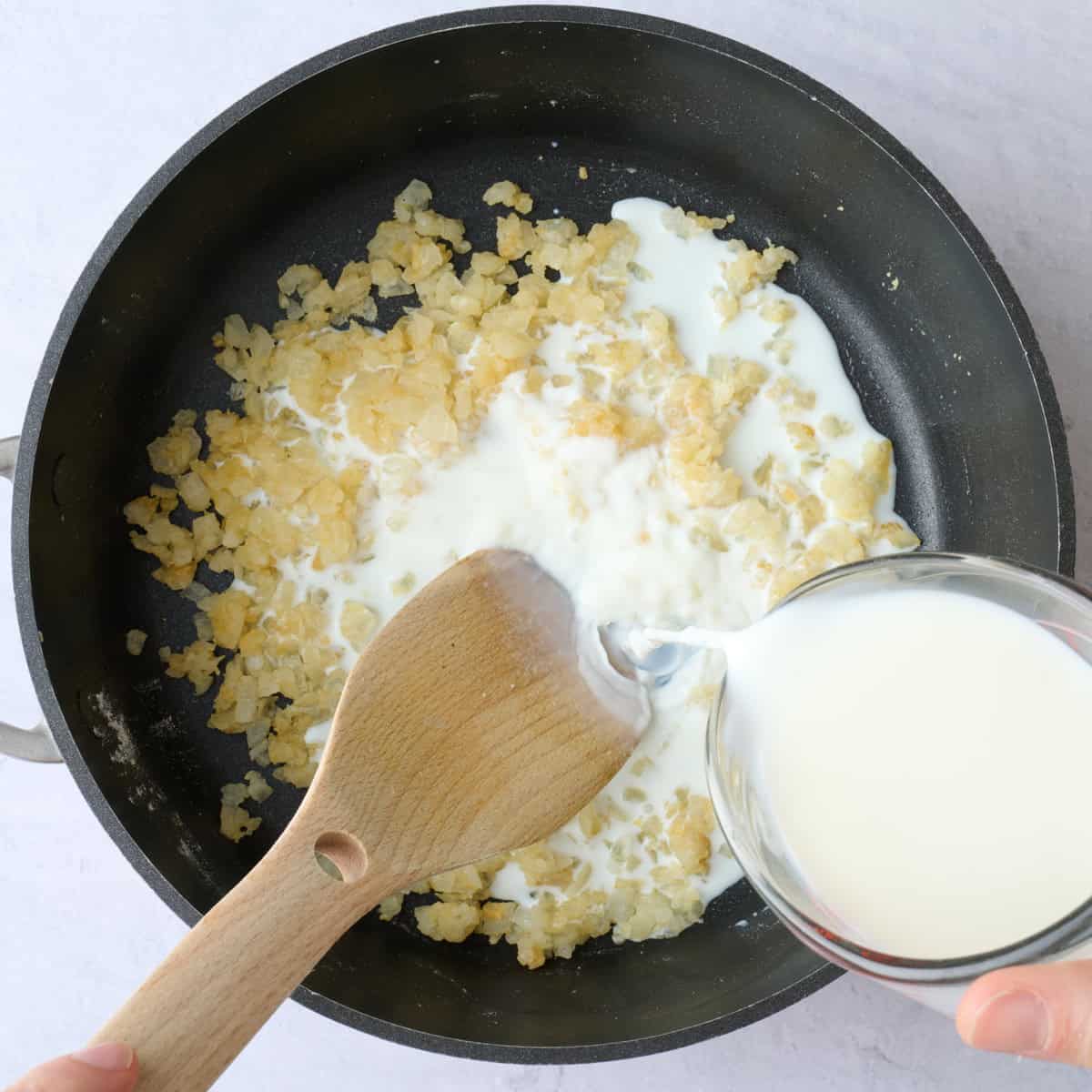 Onions with roux in a saucepan with milk being poured into it while stirring to create creamy base for casserole.
