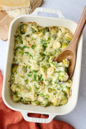 Best brussel sprout casserole in the baking dish with a serving spoon dipped inside, block of parmesan cheese nearby.
