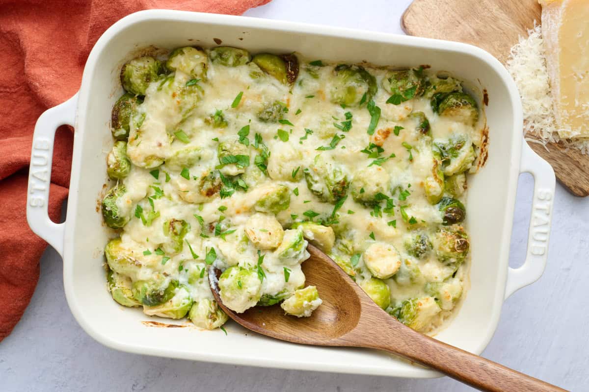 Easy brussel sprout casserole in the baking dish with a serving spoon dipped inside, block of parmesan cheese nearby.