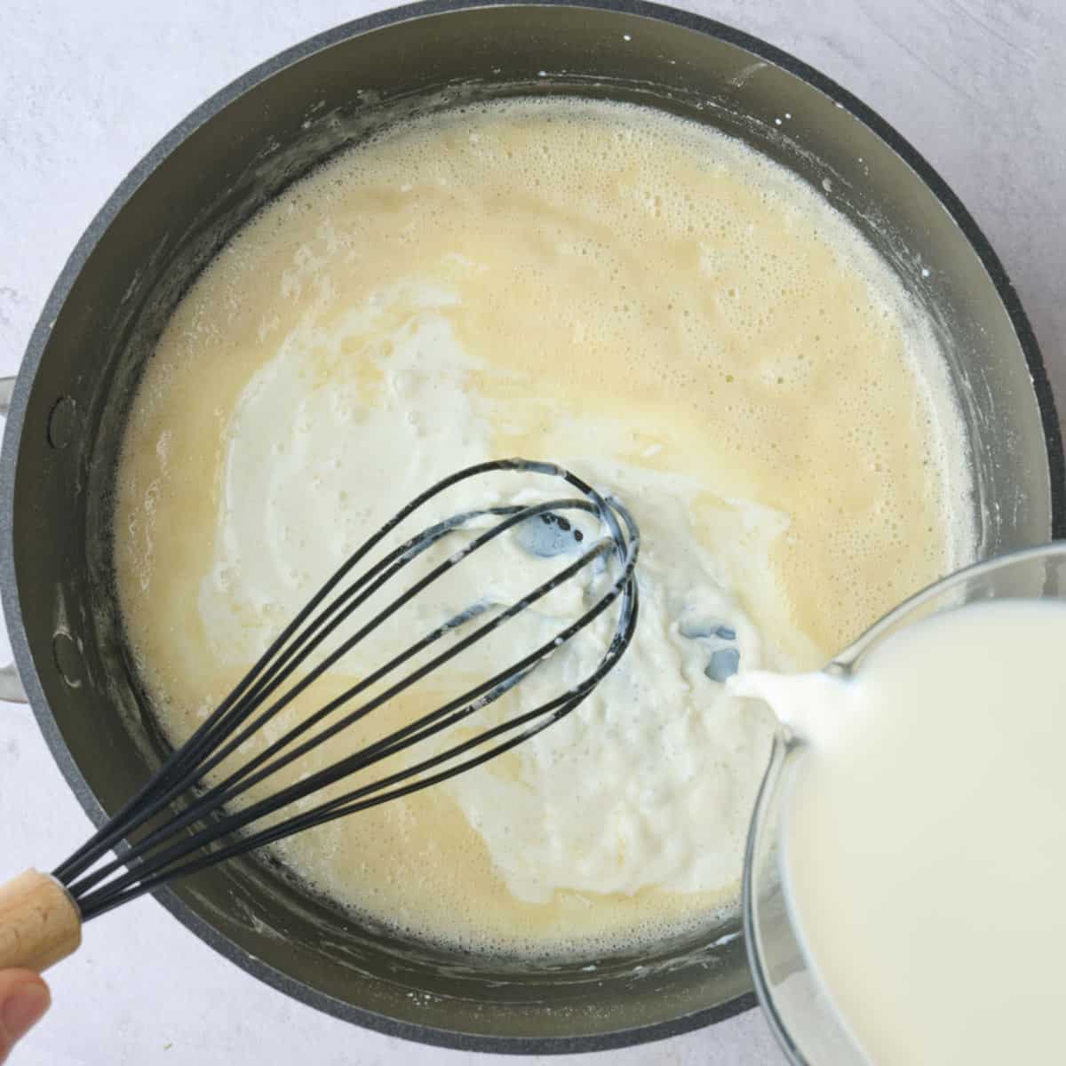 Cooked roux in a pan being whisked while milk is poured in.