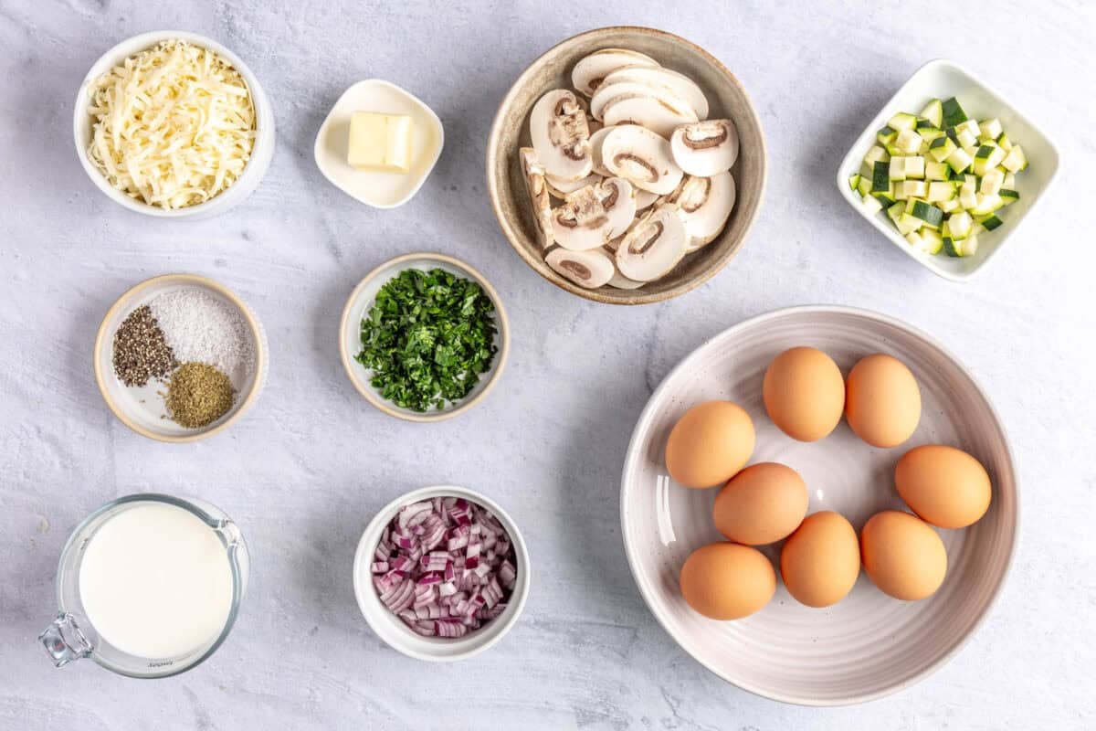 Ingredients for recipe in individual dishes for quiche recipe: shredded cheese, seasonings, milk, diced red onions, sliced mushrooms, diced zucchini, and butter.