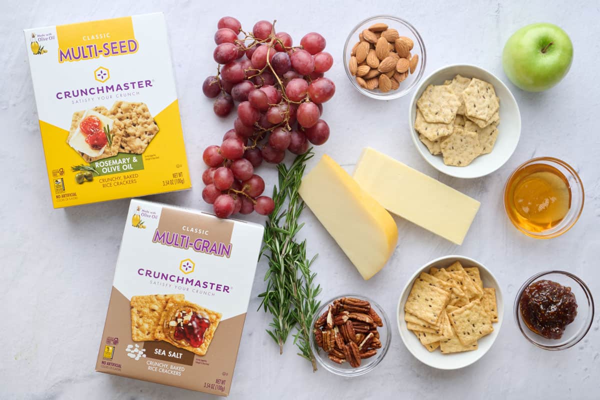 Ingredients for appetizer: Crunchmaster Rosemary & Olive Oil Crackers and Crunchmaster Sea Salt Crackers, cheese blocks, grapes, apple, mixed nuts, jam, and honey.
